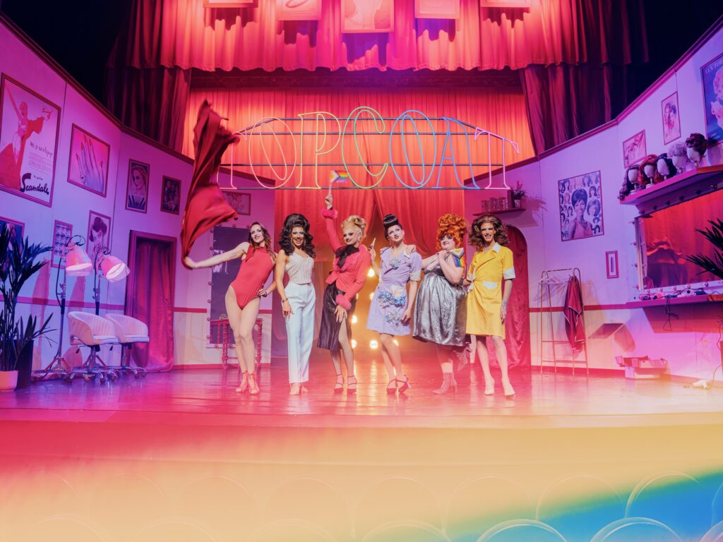 Groupe du spectacle drag Topcoat sur scène – drag queens flamboyantes et solidaires dans un Décor rétro de salon de beauté pastel inspiré des années 50
